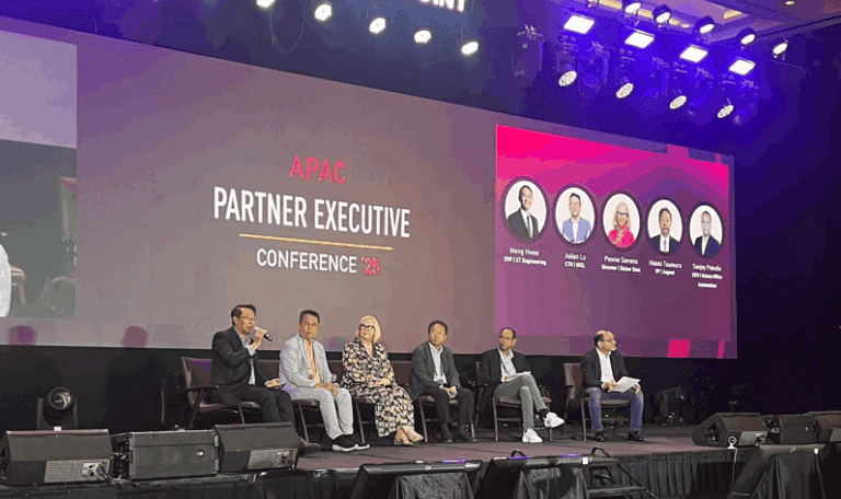 Six speakers on a conference stage participate in a panel discussion under a large screen.